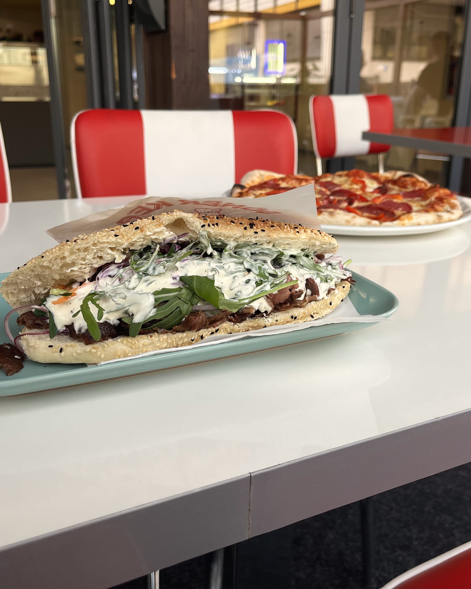 Steak-Döner-Sandwich im Sesambrot auf dem Tablett, dahinter Pizza und die rot-weißen Diner-Sitze